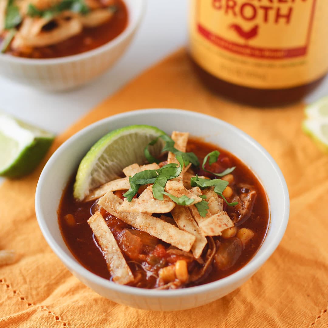 bowl of chicken tortilla soup topped with chips and lime with zoup chicken broth jar in background on table